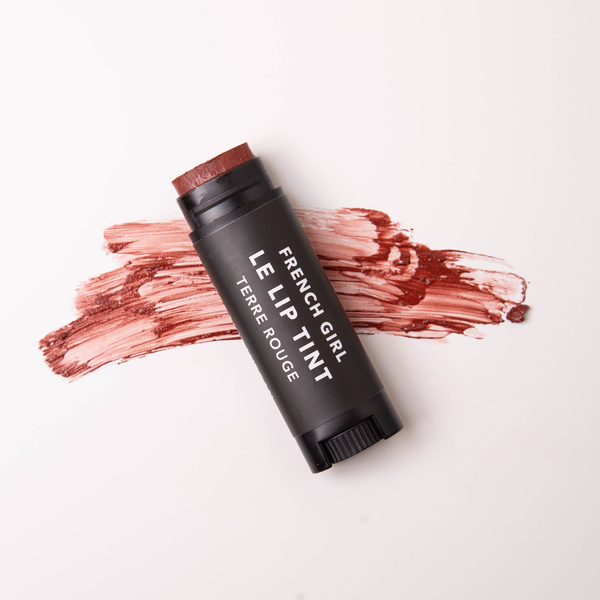 A tube of FRENCH GIRL Tinted Lip Balm in Terre Rouge rests on a white surface, with its rich reddish-brown shade swiped beneath—an organic balm that enhances your lips naturally.