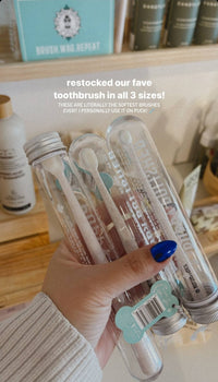 A hand with blue nail polish holds several Wag & Bright Supply Co. - Puppy Polisher Eco Toothbrushes in clear tubes. Text overlay reads, "restocked our fave toothbrush - now available in Medium size!" Shelves in the background display various bottles and products, ideal for maintaining top pet dental health.