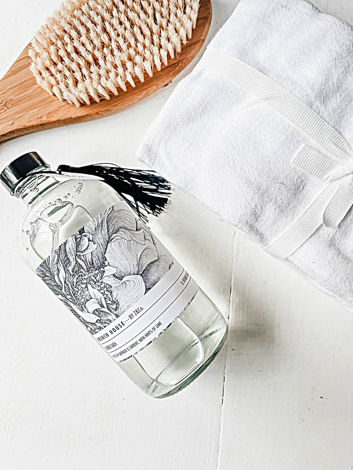 A bath brush with white bristles, a folded white towel tied with a white ribbon, and a clear glass bottle featuring the Z&Co. French House El Bosque French House Bubble Bath are neatly arranged on a white surface. The bottle contains an all-natural fragrance that promises to elevate your bathing experience.