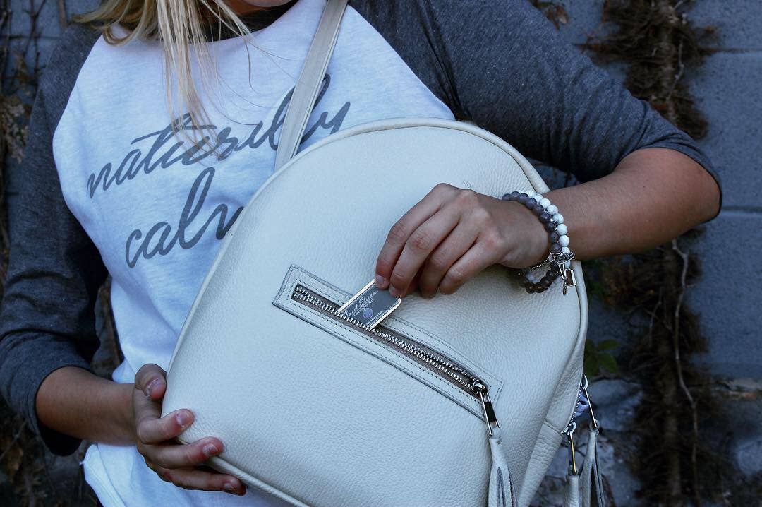 A person wearing a "naturally calm" shirt and bracelets slips an object into a white leather backpack’s front pocket. The scene highlights soothing details, echoing Sweet Streams Lavender Co.'s Lavender & Mint Lip Balm’s calming touch or its natural ingredients.