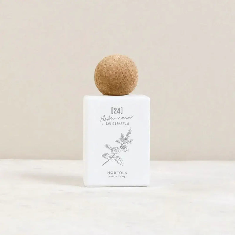 A minimalistic perfume bottle with a cork ball cap stands against a neutral background. The white rectangular bottle, adorned with a botanical illustration of fig trees, features the text "Norfolk Natural Living MidSummer Parfum 24.