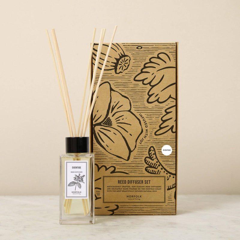 The Norfolk Natural Living Eventide Reed Diffuser Set features a glass bottle, wooden reeds, and a floral illustrated box, displayed on a light surface with a beige background for an eco-friendly home fragrance experience.