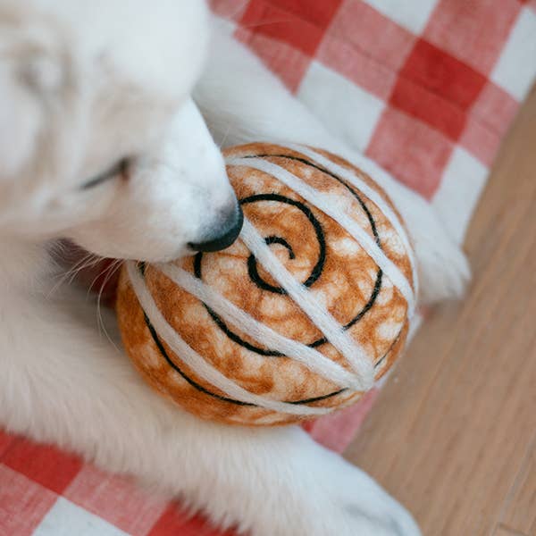 A white puppy lies on a red and white checkered blanket holding the MODERNBEAST Cinnamon Roll Wool Dog Toy by MODERNBEAST.