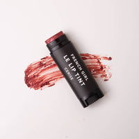 A black tube of FRENCH GIRL - Tinted Lip Balm | Cerise rests on a white surface, with a smear of the dark red vegan formula spread behind it.