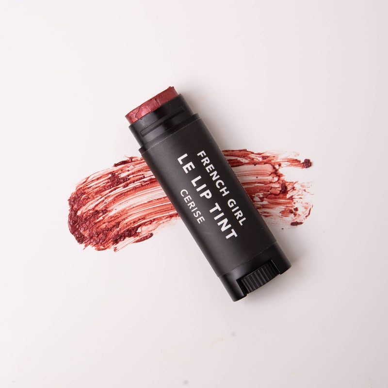 A black tube of FRENCH GIRL - Tinted Lip Balm | Cerise rests on a white surface, with a smear of the dark red vegan formula spread behind it.