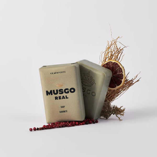 Two bars of Claus Porto 1887 Musgo Real 1887 Body Soap are shown on a white background, one upright and one tilted back, surrounded by dried twigs, a citrus slice, and red peppercorns for a nature-inspired presentation.