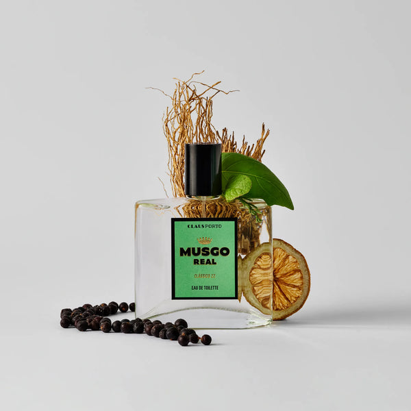A square glass bottle of Claus Porto 1887 Musgo Real Clássico 22 Eau de Toilette with a black cap is set on a white background, surrounded by dried citrus slices, green leaves, dried grass, and scattered black peppercorns.