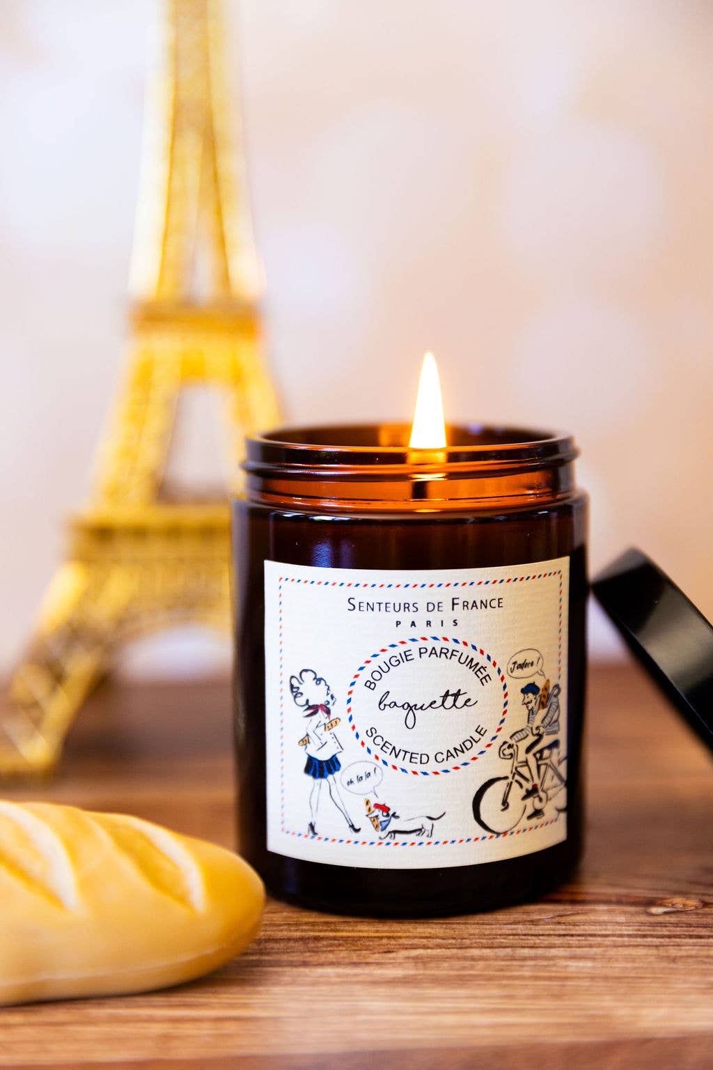 The Senteurs De France "Baguette" Candle by Senteurs De France, in an amber glass jar with French-inspired illustrations, sits on a wooden table near a baguette and Eiffel Tower model.