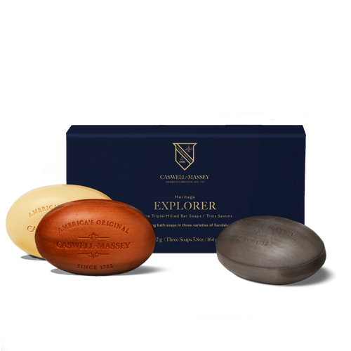 The Caswell-Massey Sandalwood Explorer Three-Soap Gift Set features three oval bar soaps—cream, brown, and dark gray—in front of a blue box. Each moisturizing, plant-based glycerin soap is embossed with "Caswell-Massey.