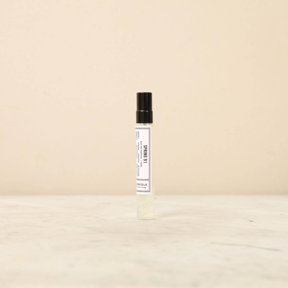 A small cylindrical spray bottle of unisex parfum is centered on a light-colored surface in front of a plain beige background. The bottle has a black cap and a white label with black text that reads "Days of Spring 91." The overall aesthetic, reminiscent of Norfolk Natural Living perfumery, is minimalistic.