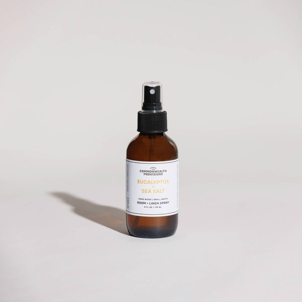 A brown Commonwealth Provisions Eucalyptus + Sea Salt Room Spray bottle stands upright on a light surface, its subtle oceanic salt notes enhancing the atmosphere against a plain, pale background.