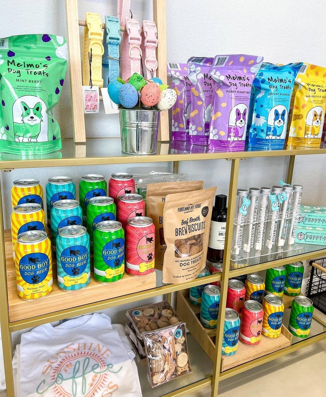 Shelves showcasing pet products include vibrant dog treats and biscuits, beverages, and accessories. The items are in bags and cans adorned with playful designs. Also visible is the Wag & Bright Supply Co.'s Puppy Polisher Eco Toothbrush (Medium) for pet dental health, alongside a shirt and various bath products. The layout is bright, inviting, and well-organized.