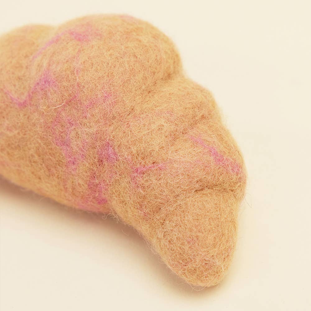 A close-up of the MODERNBEAST WOOL CROISSANT Wool Dog Toy, a brown felted wool croissant with subtle pink lines, shown on a light cream background.