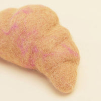 A close-up of the MODERNBEAST WOOL CROISSANT Wool Dog Toy, a brown felted wool croissant with subtle pink lines, shown on a light cream background.