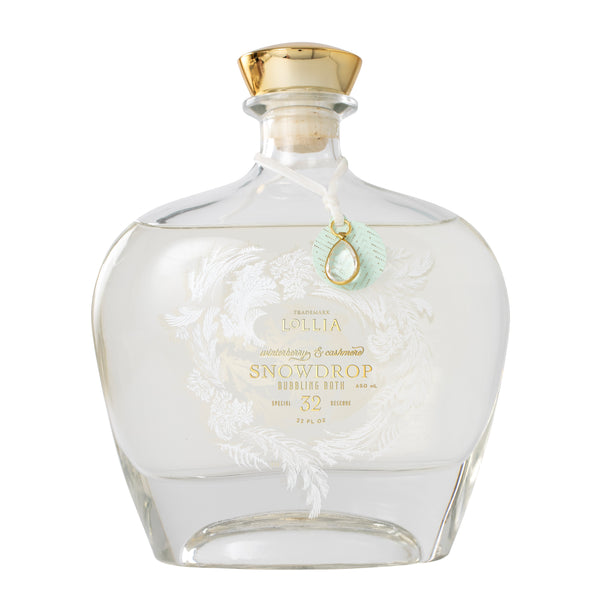 The Margot Elena Lollia Snowdrop Bubble Bath comes in a clear glass bottle with a gold-capped stopper, white floral design, blue-green pendant, and elegant gold and white lettering that showcases the Winterberry & Cashmere scent.