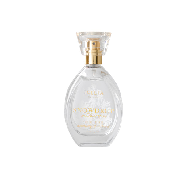 A clear glass bottle labeled "Lollia Snowdrop Eau de Parfum" by Margot Elena features a gold spray nozzle and transparent cap, with delicate white accents suggesting cashmere softness, set on a pristine white background.