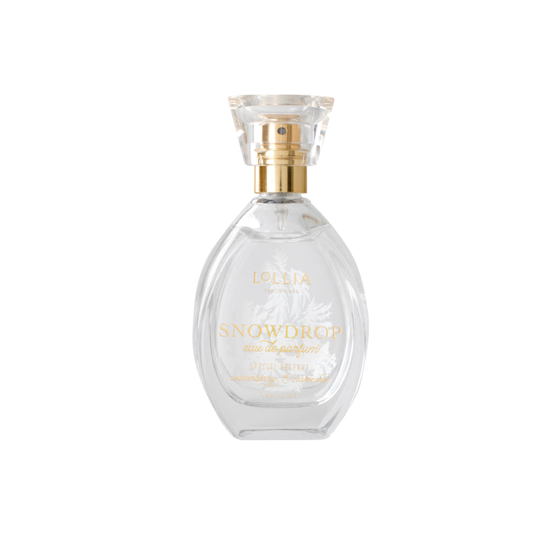 A clear glass bottle labeled "Lollia Snowdrop Eau de Parfum" by Margot Elena features a gold spray nozzle and transparent cap, with delicate white accents suggesting cashmere softness, set on a pristine white background.