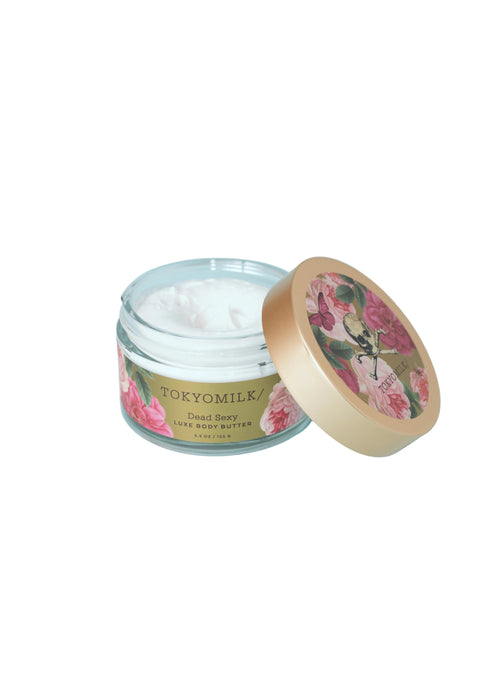 A glass jar of TokyoMilk Dead Sexy Body Butter by Margot Elena, its floral lid beside it. The partially filled luxury body butter features a label with elegant gold and pink accents.