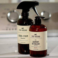 Two bottles of Fox + Hound pet laundry products, including the Natural Laundry Detergent in Basil scent, sit on a washing machine. Their natural formulas are highlighted, while blurred dials appear in the background.