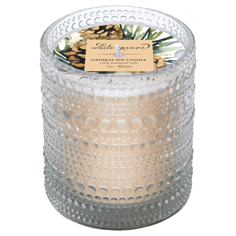 The Mangiacotti White Spruce Soy Candle 7 oz is a beige, plant-based candle in a textured clear glass holder, scented with holiday-inspired fragrance and topped with a pinecones and greenery label featuring product info.