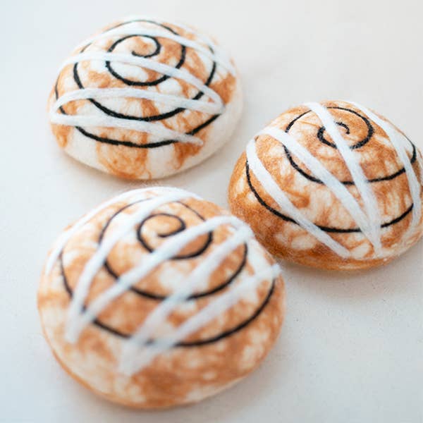 Three round MODERNBEAST Cinnamon Roll Wool Dog Toys with white icing and brown swirls rest on a light surface, highlighting the soft textures and hand-felted detail of premium New Zealand wool.