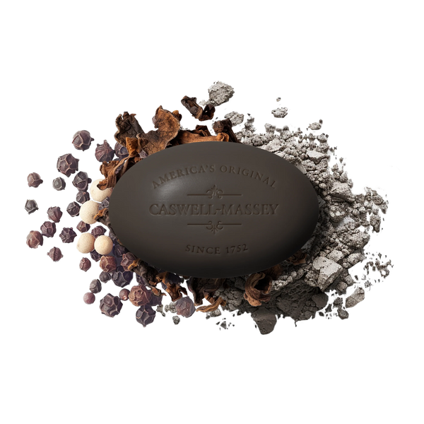 A dark brown oval Caswell-Massey Oaire Bar Soap rests on peppercorns, dried spices, and gray powder, highlighting its plant-based glycerin blend on a white background.