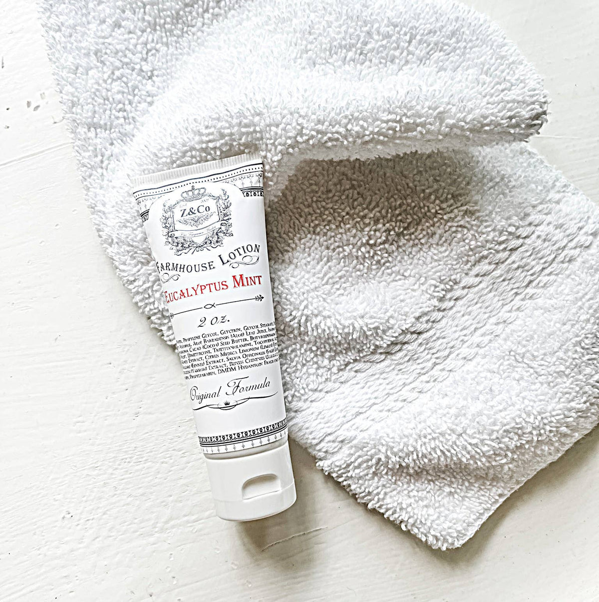 A travel-sized tube of Z&Co. Eucalyptus Mint Farmhouse Lotion (2 oz), rests on a textured white washcloth. The lotion's white packaging is adorned with intricate text and designs, offering deep hydration for your skin. Set on a smooth, white surface, the scene exudes a minimalist and clean aesthetic.
