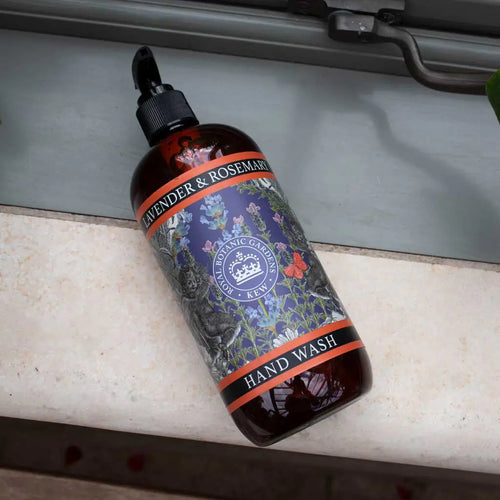 A dark amber bottle of The English Soap Co. Kew Gardens Lavender and Rosemary Hand Wash is lying on its side on a marble surface near a windowsill. The label, adorned with floral illustrations and the Kew Gardens logo, highlights ingredients like aloe vera and vitamin E. The bottle features a sleek black pump dispenser.