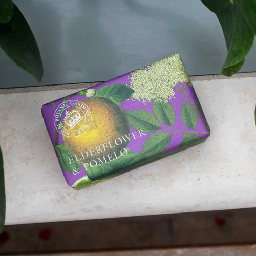 A rectangular bar of The English Soap Co. Kew Gardens Elderflower and Pomelo Soap, wrapped in purple eco-friendly packaging with pomelo, elderflower, and green leaf designs, rests on a white surface amidst lush greenery.
