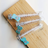 Three transparent test tubes labeled "Wag & Bright Supply Co. - Puppy Polisher Eco Toothbrush" are placed on a wooden surface. Featuring silver caps and blue tags, these innovative tubes hold the medium-sized toothbrush made by Wag & Bright Supply Co., crafted from wheat straw bioplastic to sustainably enhance pet dental health.