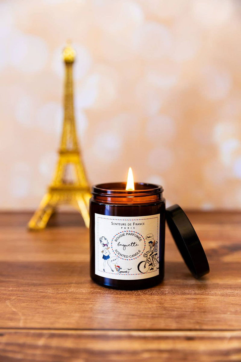 The Senteurs De France "Baguette" Candle by Senteurs De France, in a brown glass jar with its lid off, sits on a wooden surface, while a small Eiffel Tower model is blurred in the background.