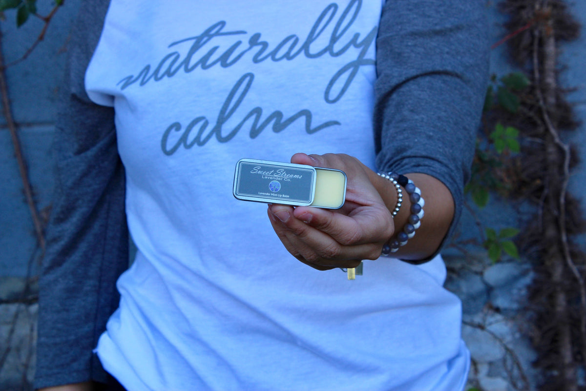 Someone in a gray and white long-sleeve shirt with "naturally calm" holds a small rectangular tin labeled "Lemon Lavender." This Sweet Streams Lavender Co. - Lavender & Mint Lip Balm, made with natural ingredients, is set against a stone wall and lush foliage.