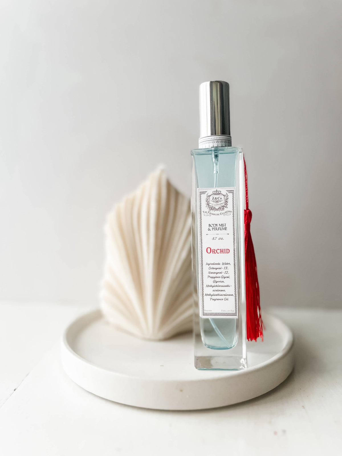 A clear glass bottle of Z&Co. Farmhouse Body Mist - Orchid with a silver cap and red tassel sits on a round white tray, with an abstract folded white object in the background, reflecting the Z&Co. exotic fragrance experience.