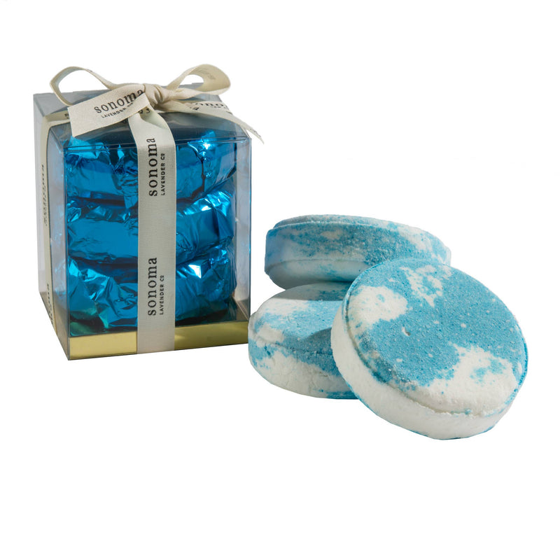 The "Sonoma" labeled translucent gift box, adorned with a beige ribbon, holds four blue and white Sonoma Lavender Ocean Aire shower steamers. Alongside the box are two additional steamers that capture the calming fragrance of Sonoma Lavender.