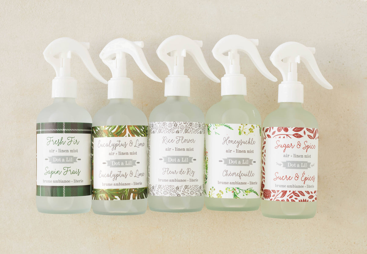 Five bottles of Dot & Lil Linen + Air Mist are displayed, each with a unique label: Fresh Fir, Eucalyptus & Lime, Rice Flower, Honeysuckle, and Sugar & Spices. All are vegan room sprays from Dot & Lil.