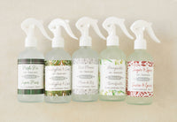 Five bottles of Dot & Lil Linen + Air Mist are displayed, each with a unique label: Fresh Fir, Eucalyptus & Lime, Rice Flower, Honeysuckle, and Sugar & Spices. All are vegan room sprays from Dot & Lil.