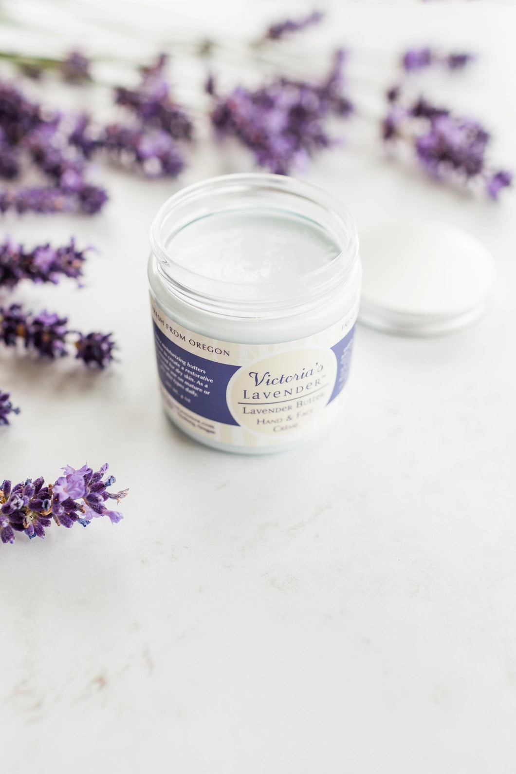 An open jar of Victoria's Lavender - Lavender Butter Face & Hands - Travel Size is placed on a white surface, surrounded by scattered sprigs of fresh lavender. The label reads "Victoria's Lavender, Hand and Body Cream." Renowned for its non-greasy formula, this luxurious lavender buttercream has a travel size lid lying beside it.