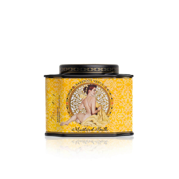 A rectangular tin from Barefoot Venus features a seated woman modestly draped in fabric against a yellow background with ornate patterns. The product name "Barefoot Venus Mustard Bath Soak - 100% Natural" is visible. It's cruelty-free and has a black rim and lid.