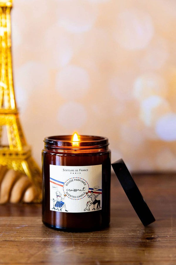 The Senteurs De France "Croissant" candle by Senteurs De France glows on a wooden surface, its French-themed label and black lid nearby with an Eiffel Tower decor accentuating the Parisian ambiance in the background.