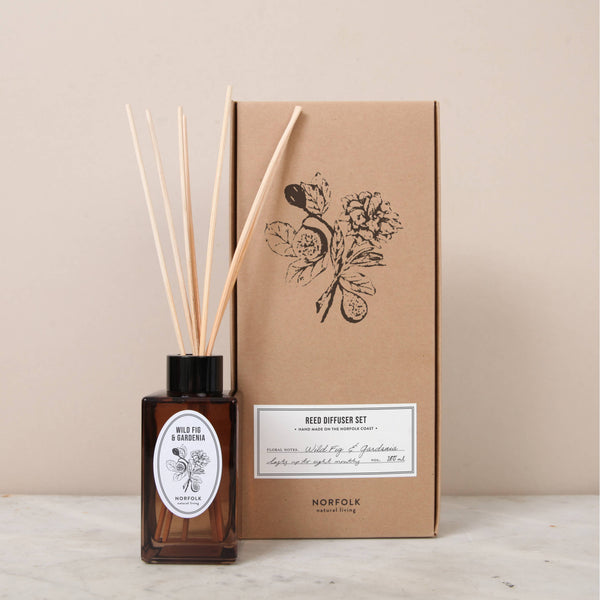 The Norfolk Natural Living Wild Fig & Gardenia Reed Diffuser Set (100ml) features a brown glass diffuser with wooden reeds beside its eco-friendly, botanical-illustrated kraft box, both elegantly displayed on a neutral surface.