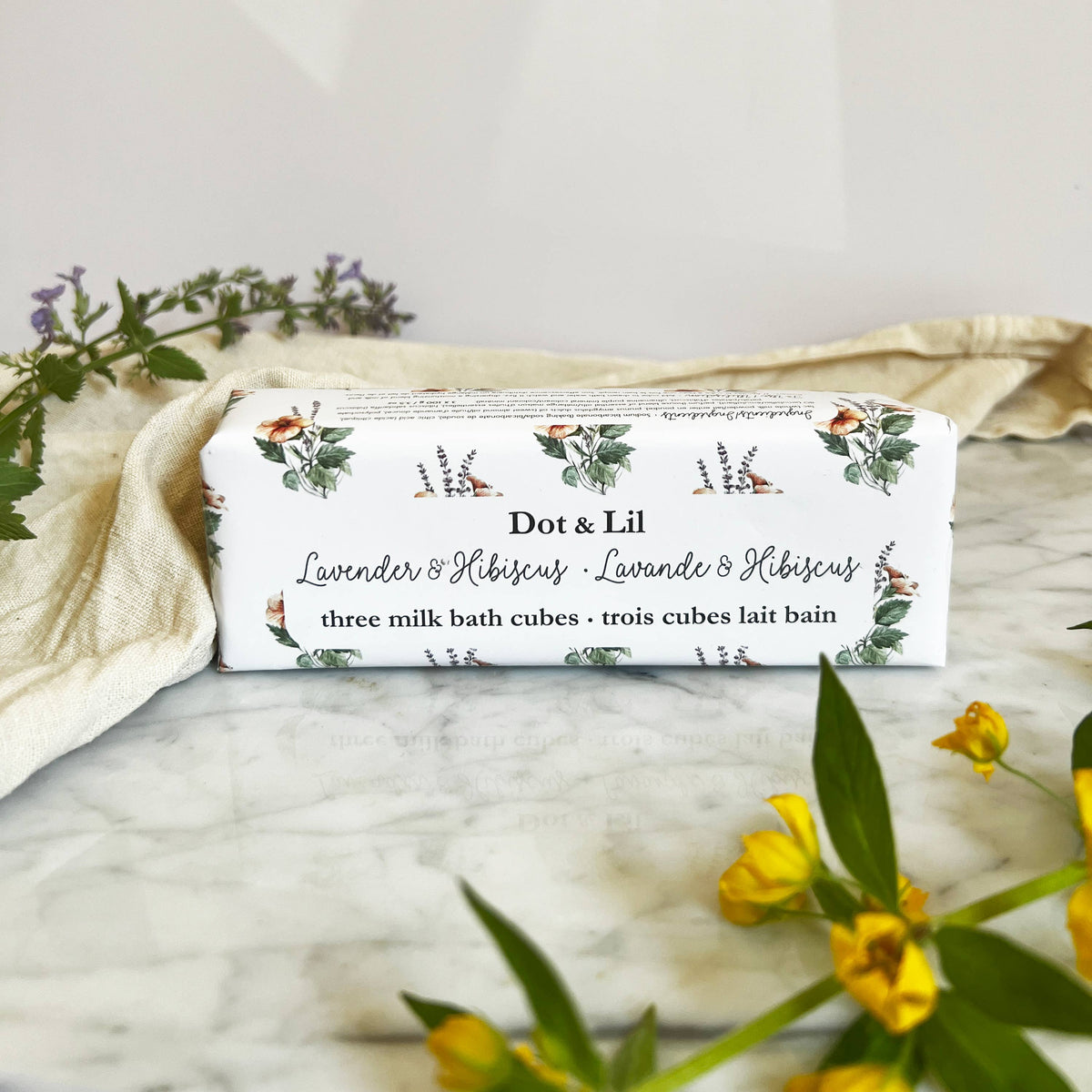 A rectangular box labeled "Dot & Lil Lavender & Hibiscus Milk Bath Cubes Trio Gift Set" rests on marble, surrounded by yellow and purple flowers and a cream cloth, capturing the luxurious bath experience offered by Dot & Lil.