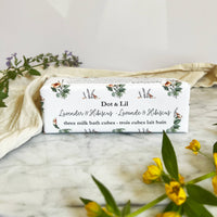 A rectangular box labeled "Dot & Lil Lavender & Hibiscus Milk Bath Cubes Trio Gift Set" rests on marble, surrounded by yellow and purple flowers and a cream cloth, capturing the luxurious bath experience offered by Dot & Lil.