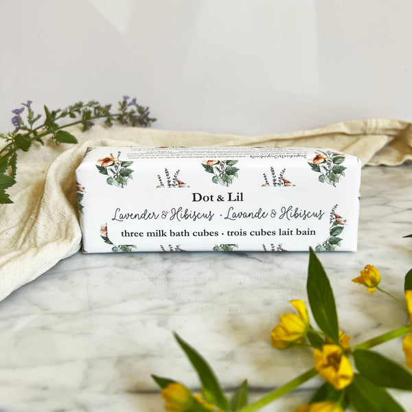 A rectangular box labeled "Dot & Lil Lavender & Hibiscus Milk Bath Cubes Trio Gift Set" rests on marble, surrounded by yellow and purple flowers and a cream cloth, capturing the luxurious bath experience offered by Dot & Lil.