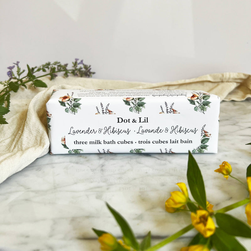 A rectangular box labeled "Dot & Lil Lavender & Hibiscus Milk Bath Cubes Trio Gift Set" rests on marble, surrounded by yellow and purple flowers and a cream cloth, capturing the luxurious bath experience offered by Dot & Lil.
