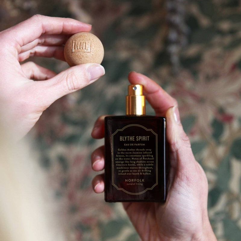 A person holds a Norfolk Natural Living Blythe Spirit Eau de Parfum - 50ml bottle in one hand and its round wooden cap in the other, highlighting the fragrance against a blurred, leafy background.