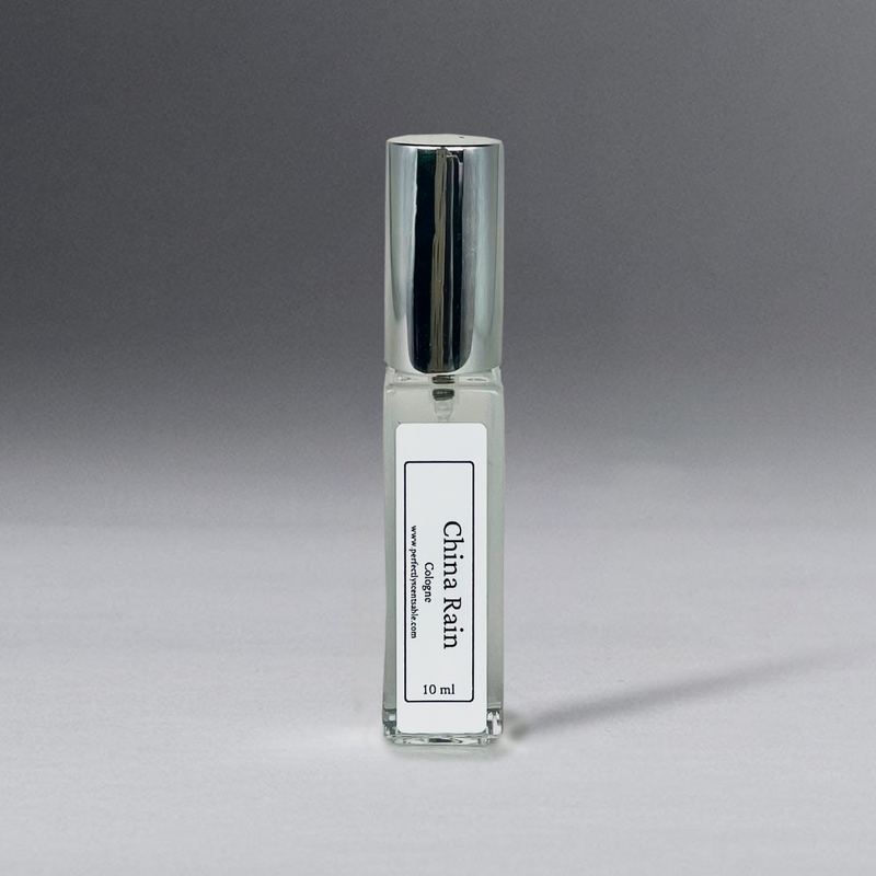 A small, transparent glass bottle with a metallic cap, containing a clear liquid labeled "China Rain" in vertical text. Infused with the subtle musk of white lilies, the Perfectly Scentsable China Rain Perfume Oil - 10ml Cologne Spray is elegantly showcased against a gradient gray background.