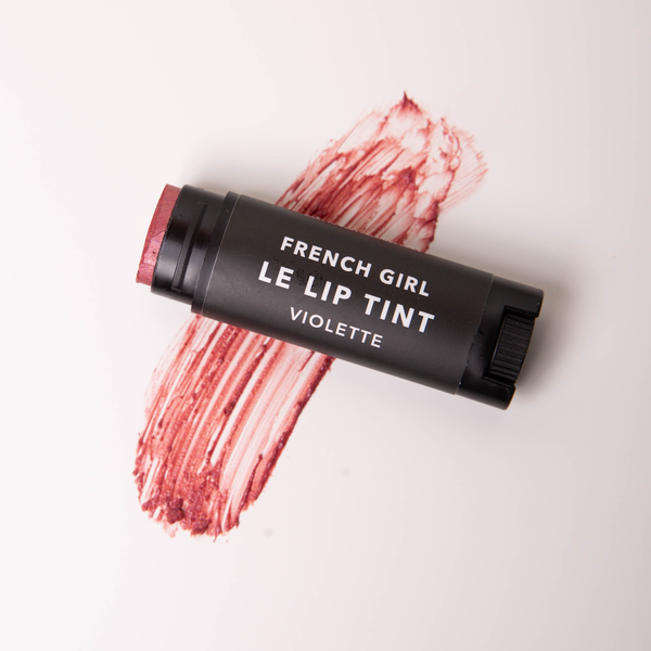 A tube of FRENCH GIRL Tinted Lip Balm | Violette rests on a white surface, with a swipe of mauve color beneath it.