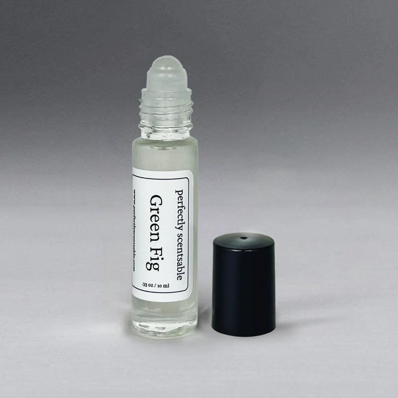 A small, clear glass bottle with a rollerball applicator labeled "Perfectly Scentsable Green Fig Perfume Oil Roll-On." The bottle contains liquid and has a black cap placed beside it on a gray background.