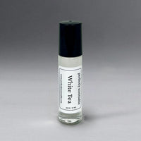 A small glass roll-on bottle with a black cap labeled "Perfectly Scentsable White Tea Perfume Oil - Roll-On" by Perfectly Scentsable. The label includes additional text and a website URL. The bottle contains a clear liquid infused with hints of fragrance, musk, and amber, set against a gradient gray background.