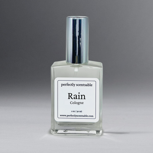 A square, clear glass bottle of cologne labeled "Perfectly Scentsable Rain Perfume Oil - 1oz Cologne Spray" on a white label with black text. The label also reads "Perfectly Scentsable" at the top and "1 oz / 30 ml" at the bottom, along with a website URL. The bottle has a shiny, silver cylindrical cap and features a gender-neutral aroma.